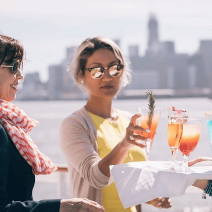 Two women standing outdoors with a city skyline blurred in the background, as one of them takes a colorful drink from a tray.
