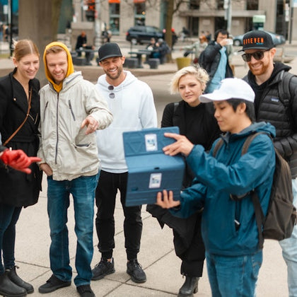 A group of people in casual clothing gathered outdoors, smiling and looking at a blue box held by a person in a blue jacket.
