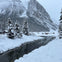 Snow-covered landscape with a river, tall pine trees, and distant people on a frozen lake. A mountain range is in the background.