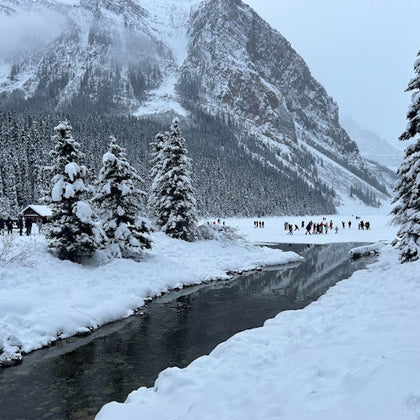 Snow-covered landscape with a river, tall pine trees, and distant people on a frozen lake. A mountain range is in the background.