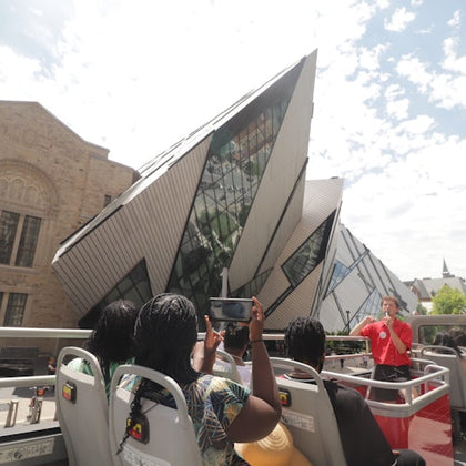 The tour guide is talking on the mic on the upper level of an open-top double-decker tour bus, passing the Royal Ontario Muse