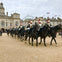 A ceremonial procession of mounted guards in uniform passing in front of a historic building with flags and a Ferris wheel in the background.
