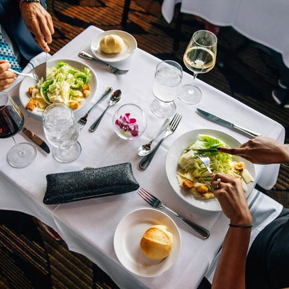 Two people having a meal at a restaurant table with salads, bread rolls, water, wine, and silverware on a white tablecloth.
