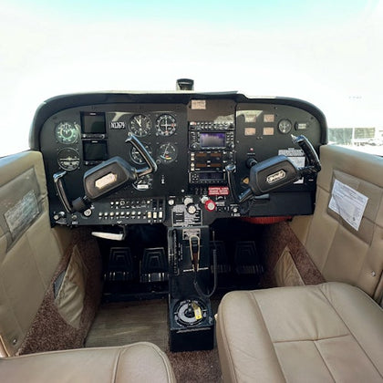 Cockpit of a small aircraft featuring dual control yokes, various instrument gauges, switches, and beige seats.