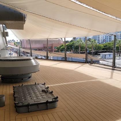 A ship's deck with covered shade, a hatch, and mechanical equipment; buildings and trees are visible in the background.