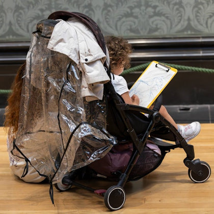 A child in a stroller draws on a clipboard, covered by a transparent rain cover.
