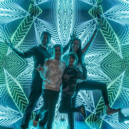 Four people are posing energetically in front of an illuminated background with intricate, symmetrical neon patterns.