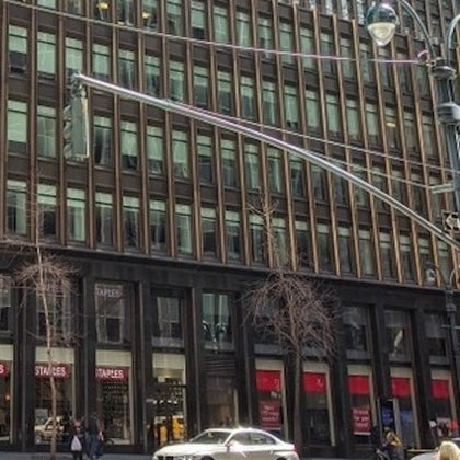 Street view of a building with many windows, a Staples store on the ground level, and a car passing by in the foreground.