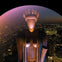 A stylized statue with geometric features and headdress stands illuminated against a backdrop of a city at night.