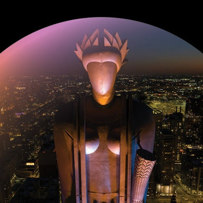 A stylized statue with geometric features and headdress stands illuminated against a backdrop of a city at night.