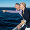 A man and a woman smiling and leaning over a boat railing, with the ocean and a clear blue sky in the background.