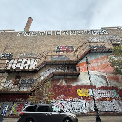 Brick building with extensive graffiti, metal fire escapes, and a parked minivan in front. Overcast sky above.