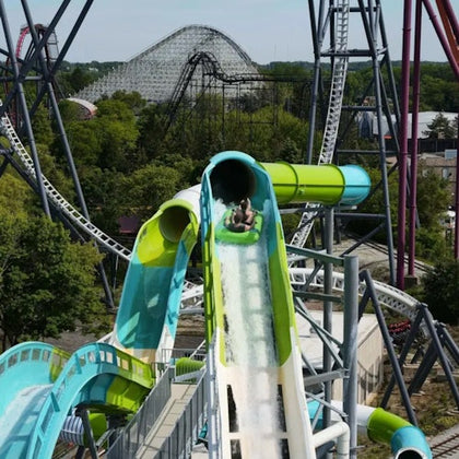 A person sliding down a water slide with roller coasters and trees in the background on a sunny day.