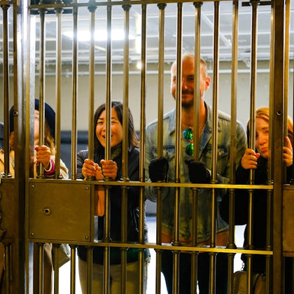 A group of people standing behind a metal gate, smiling and holding the bars. The background shows an indoor setting.
