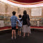 Three people stand in front of a large, illuminated manuscript display in a museum setting.