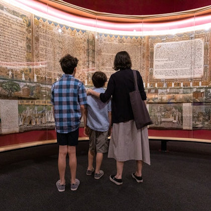 Three people stand in front of a large, illuminated manuscript display in a museum setting.
