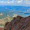 A view from a rocky mountain summit overlooking a vast landscape of forests, valleys, and distant hills under a cloudy sky.