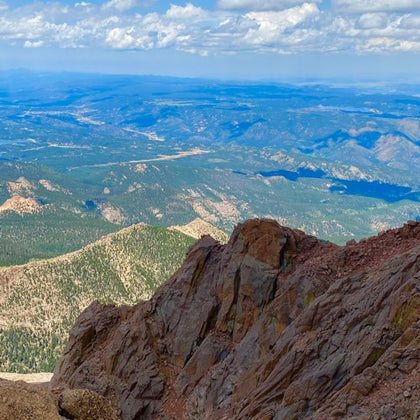 A view from a rocky mountain summit overlooking a vast landscape of forests, valleys, and distant hills under a cloudy sky.