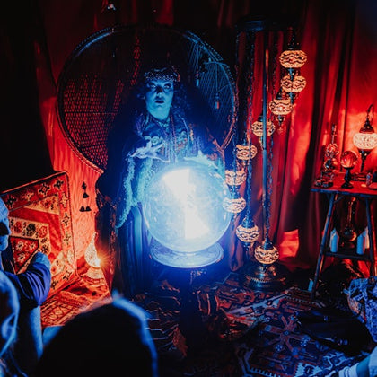 A person in dramatic lighting gazes into a glowing crystal ball in a dimly lit room with ornate decor and hanging lamps.
