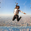 Person jumping inside a glass observation deck, smiling with arms outstretched, with a cityscape view in the background.