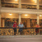 Three people stand on an ornate balcony inside a historic building with murals and flags in the background.