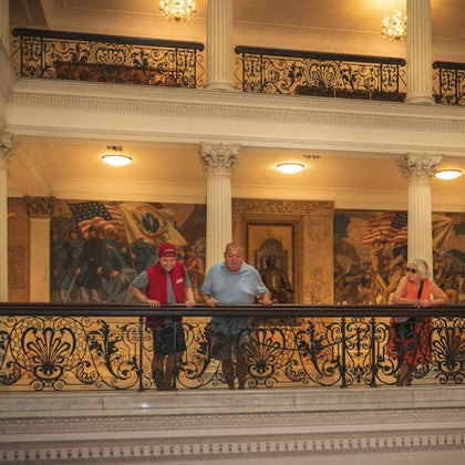 Three people stand on an ornate balcony inside a historic building with murals and flags in the background.
