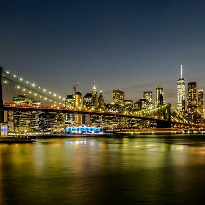 City skyline at night with a lit suspension bridge over a river and illuminated buildings in the background.