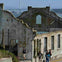 Ruins of a building with people walking nearby, a bridge and water visible in the background.