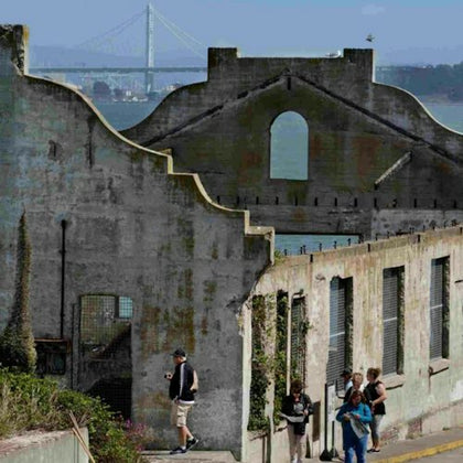 Ruins of a building with people walking nearby, a bridge and water visible in the background.
