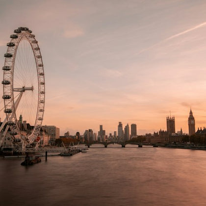 A large ferris wheel beside a river, with a bridge and city skyline in the background at sunset.