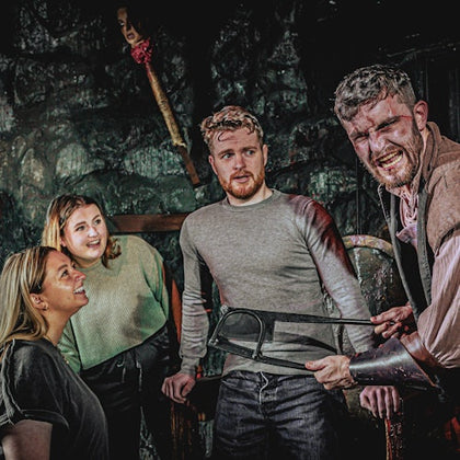 Three people look at a man holding a saw with a grimace in a dimly lit, eerie room decorated with a severed head on a pike.