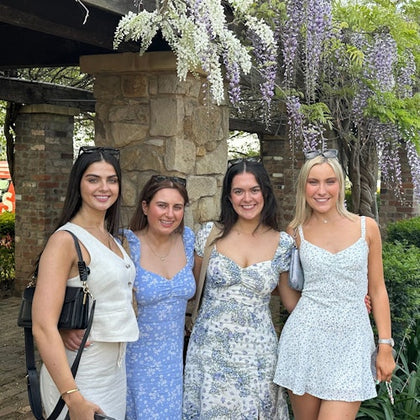 4 women standing in front of a walkway covered in purple flowers. The Dave's bus can be seen in the background.