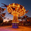 An illuminated carousel swing ride spins with riders at dusk, surrounded by palm trees and colorful lights.