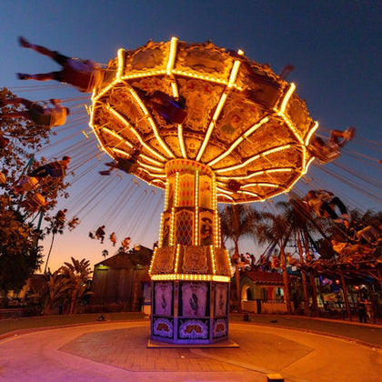 An illuminated carousel swing ride spins with riders at dusk, surrounded by palm trees and colorful lights.