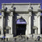 People walking in front of a large classical-style building with columns and an arched entrance, featuring a banner above the doorway.