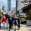 A group of people posing and smiling outdoors in a cityscape, with tall buildings, trees, and a clear blue sky in the background.