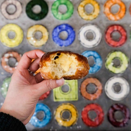 Hand holding a bitten glazed doughnut, in front of a background of various colorful doughnuts arranged in rows.