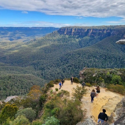 Several people stand on a cliff edge overlooking a vast, forested valley and distant mountains under a partly cloudy sky.