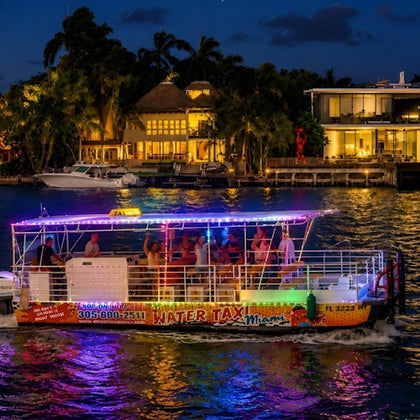 A lit water taxi with passengers cruises along a waterfront at night, with illuminated buildings and trees in the background.