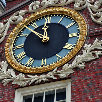 Large decorative clock with Roman numerals, gold accents, and black face mounted on a red brick building above a window.
