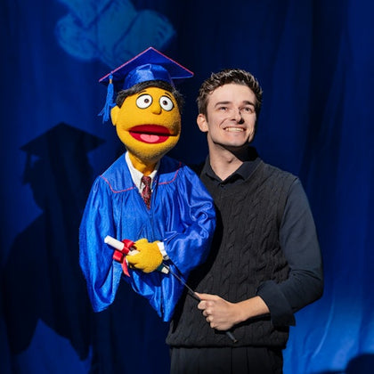 A person holding a puppet wearing a graduation cap and gown, standing against a blue background with a shadow.