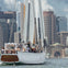 A sailboat named "Adirondack IV" full of passengers glides on water with a city skyline of modern buildings in the background.