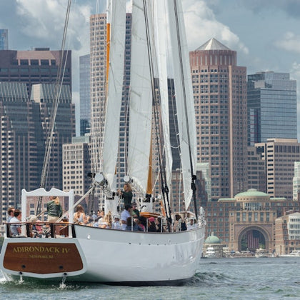 A sailboat named "Adirondack IV" full of passengers glides on water with a city skyline of modern buildings in the background.