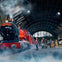 A red steam engine labeled "Hogwarts Express" at a train station with people handling luggage and a clock on the wall.