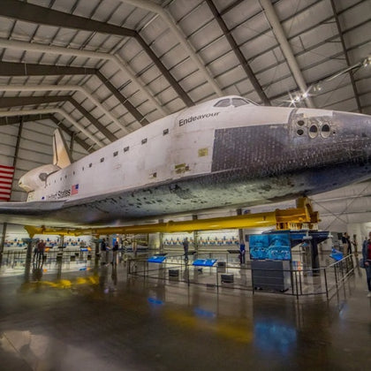 Space Shuttle Endeavour displayed inside a large museum hangar, surrounded by informational exhibits and an American flag on the wall.