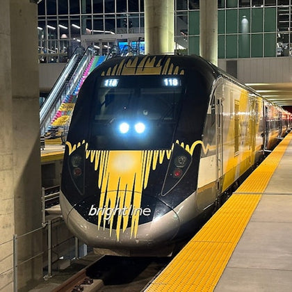 A Brightline train stopped at a station platform with yellow safety lines and an escalator visible in the background.