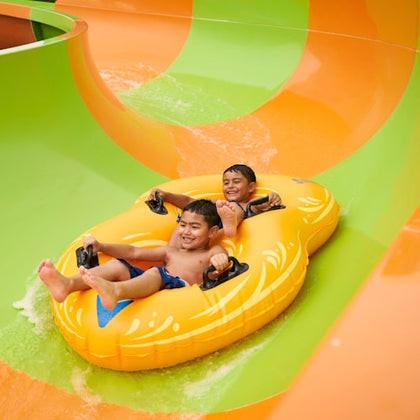 Two children smiling while riding a yellow inflatable raft down a water slide with orange and green sections.