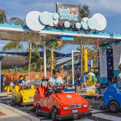 Children driving small Lego-style cars through a playful car wash attraction with bubbles, palm trees, and a sunny sky.