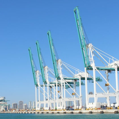 A row of large green and white cranes at a port with a city skyline in the background under a clear blue sky.