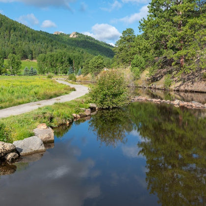 A peaceful scene with a dirt path beside a calm stream, surrounded by lush greenery and forested hills under a blue sky.